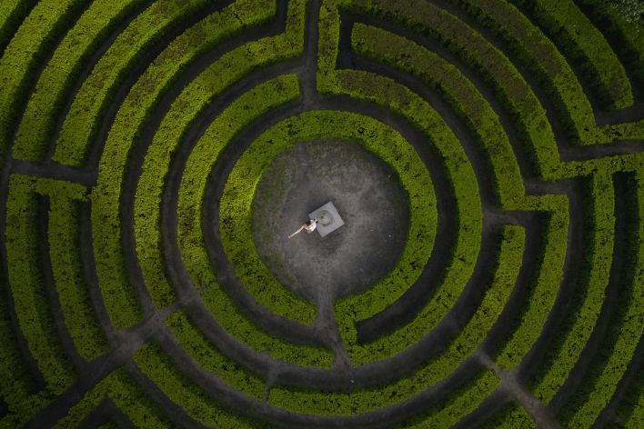 Drone shot of a woman standing in the center of a lush green hedge maze.